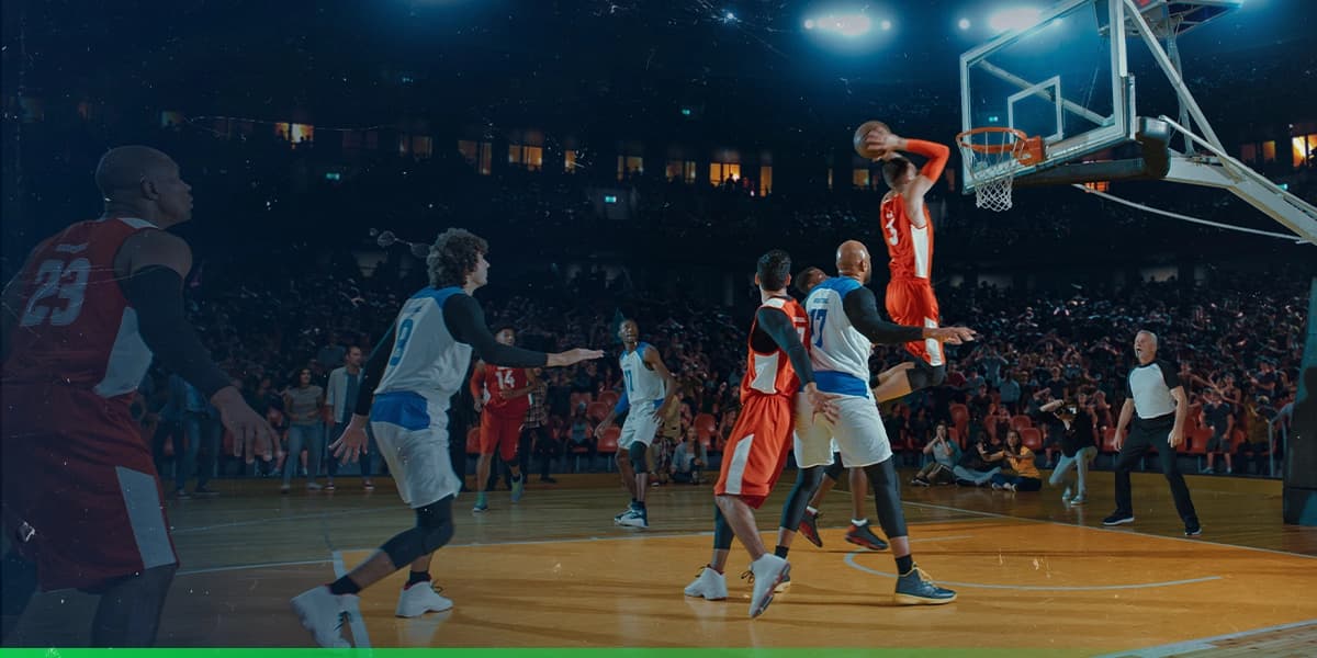 Imagem de um jogo de basquete entre duas equipes onde a equipe de time de vermelho tem um jogador prestes a dar uma enterrada e marcar ponto