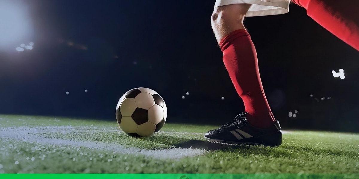 Jogador de futebol prestes a chutar uma bola em campo iluminado durante a noite; ele veste meião vermelho, chuteira preta e uniforme branco, com foco nos pés e na bola posicionada sobre a grama.